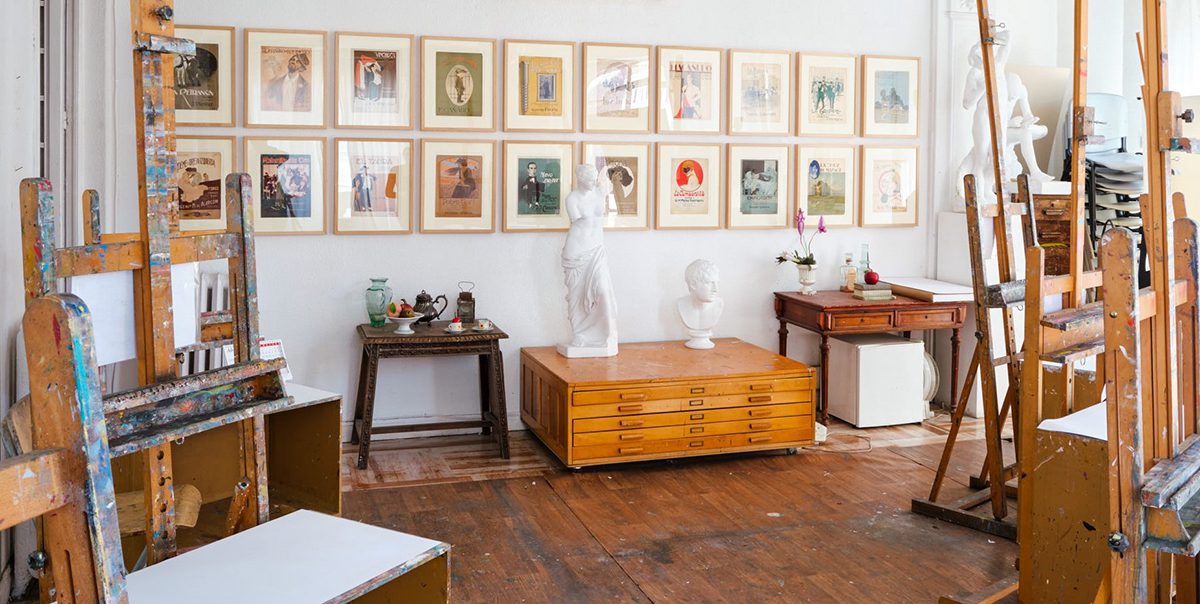 Interior de un estudio de arte luminoso con caballetes, obras enmarcadas en la pared y esculturas sobre muebles de madera.